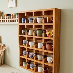 Può includere: Un portabicchieri in legno con più scomparti, ognuno dei quali contiene una tazza di ceramica diversa. Le tazze variano per colore e design, tra cui bianco, blu e marrone. Il portabicchieri è montato su una parete verde chiaro.