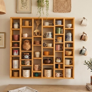 Pu&ograve; includere: Una mensola in legno con pi&ugrave; scomparti, piena di tazze, tazzine e oggetti decorativi. La mensola &egrave; montata su una parete beige, con tazze aggiuntive appese a ganci. Gli articoli sono di vari colori e design.