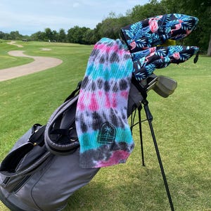May include: A gray golf bag with a black and white tie-dye towel hanging from it. The bag is on a golf course with green grass and a winding path. The golf clubs have pink flamingo headcovers.