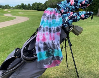 May include: A gray golf bag with a black and white tie-dye towel hanging from it. The bag is on a golf course with green grass and a winding path. The golf clubs have pink flamingo headcovers.