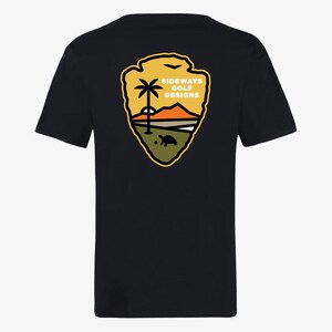 Peut inclure: T-shirt noir avec un graphique jaune, orange et vert d'un badge de garde forestier avec un palmier, un coucher de soleil et une tortue. Le texte "Sideways Golf Designs" est sur le badge.