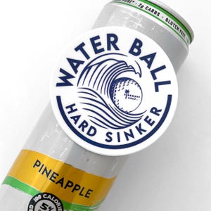 May include: White round sticker with blue text and a golf ball graphic. The text reads "Water Ball Hard Sinker". The sticker is on a white can with the text "Pineapple" in yellow.
