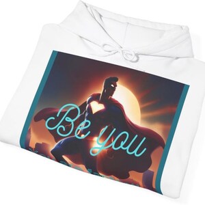 Könnte beinhalten: Weißer Kapuzenpullover mit einer Grafik eines Superhelden, einem herzförmigen Emblem und einem Sonnenuntergangshintergrund. Der Schriftzug "Be you" ist türkis. Der Kapuzenpullover hat eine Kapuze mit Kordelzug.