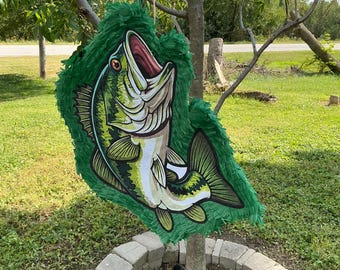 May include: A fish-shaped piñata featuring a green and white largemouth bass illustration with an open mouth, surrounded by green fringed paper accents. The piñata is suspended from a tree branch by a thin rope, displaying detailed scales and fins.