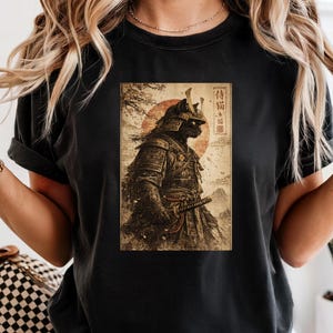 May include: Black t-shirt featuring a vintage-style graphic of a samurai cat in full armor, with a katana. The design includes a red sun and Japanese text. The shirt is a classic crew neck.
