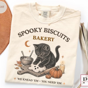 Peut inclure: Un t-shirt ivoire avec un motif représentant un chaton noir pétrissant de la pâte sur une planche en bois. Le texte indique SPOOKY BISCUITS BAKERY et WE KNEAD 'EM YOU NEED 'EM. Le design inclut un bol, un fouet, des citrouilles, une bougie et un croissant de lune.