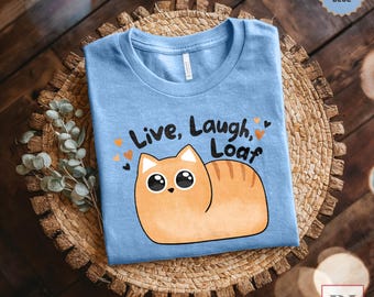 Op de afbeelding: Een lichtblauw t-shirt met een cartoon oranje kat en de tekst "Live, Laugh, Loaf".