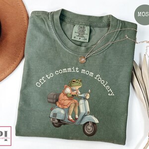 Peut inclure: T-shirt vert mousse avec l'inscription "Off to commit mom foolery" et une grenouille sur un scooter bleu clair. La grenouille porte une robe. Un collier argenté est drapé sur le t-shirt. Marque Comfort Colors.