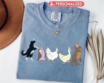 May include: A light blue Comfort Colors t-shirt featuring a line of cartoon animals wearing party hats. The animals include a black and white cat, an owl, a chicken, a duck, and a bear. A gold necklace is draped across the shirt.