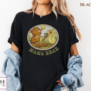 May include: Black t-shirt featuring a vintage-style graphic with a brown bear, yellow duck, and butterfly. The graphic is inside a circle with the words "MAMA BEAR" in green. The shirt is a casual, comfortable style.