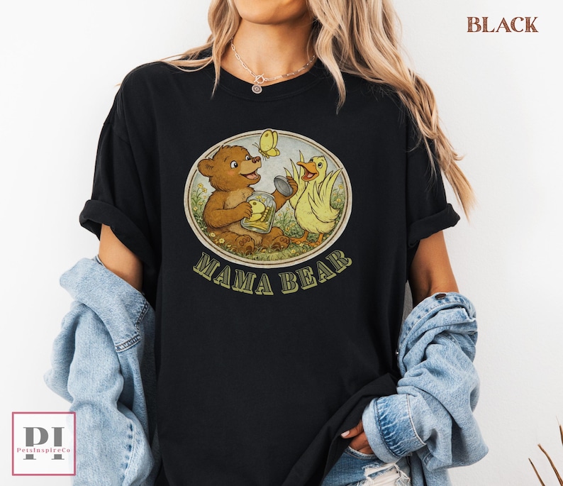 May include: Black t-shirt featuring a vintage-style graphic with a brown bear, yellow duck, and butterfly. The graphic is inside a circle with the words "MAMA BEAR" in green. The shirt is a casual, comfortable style.