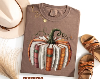 Könnte beinhalten: Espressofarbenes T-Shirt mit einem Kürbisdesign aus Büchern. Die Grafik hat einen grünen Stiel und orange, rote und braune Buchrücken. Eine goldene Halskette und braune Sandalen sind ebenfalls im Bild. Das T-Shirt ist mit "XL-TG-EG" gekennzeichnet.