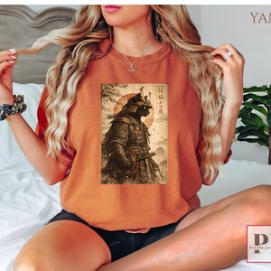 May include: Burnt orange t-shirt featuring a detailed graphic of a samurai warrior with a katana, set against a rising sun and Japanese text. The design is printed on a comfortable, casual tee, perfect for fans of Japanese culture and history.