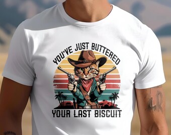 May include: White t-shirt featuring a vintage-style graphic of a cat wearing a cowboy hat and holding two guns. The text "You've Just Buttered Your Last Biscuit" is printed below the cat.
