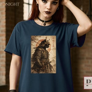 May include: Navy blue t-shirt featuring a detailed illustration of a samurai cat with a red sun in the background. The shirt has the word "MIDNIGHT" printed at the top left corner. The design is in a vintage style.