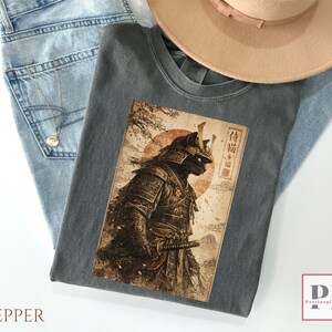 May include: A gray t-shirt featuring a detailed illustration of a black cat in samurai armor, set against a backdrop of a red sun and Japanese text. The shirt is folded on top of blue jeans and a beige hat.