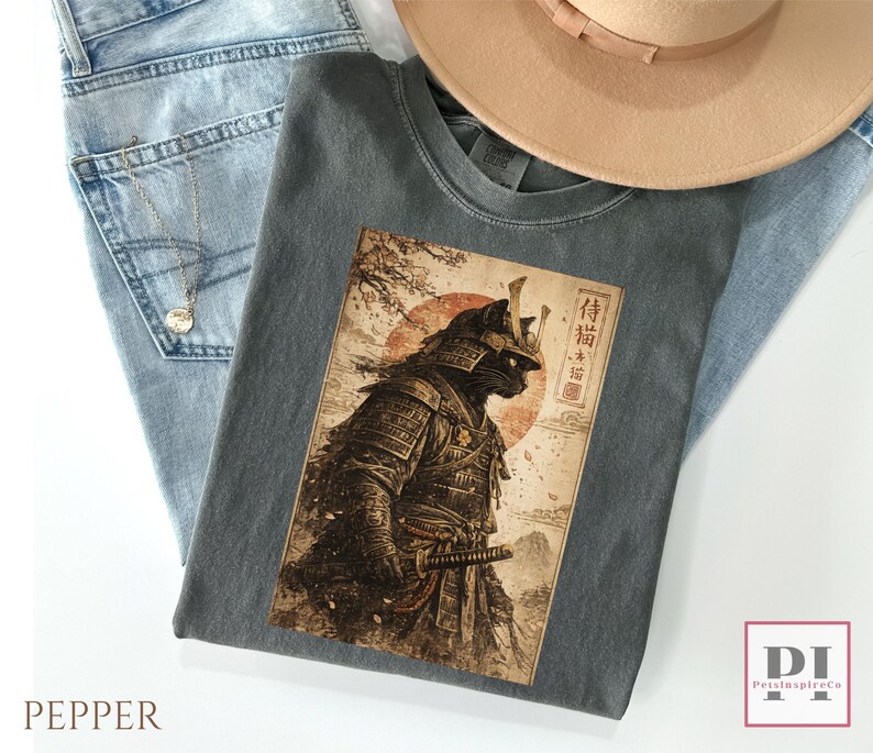 May include: A gray t-shirt featuring a detailed illustration of a black cat in samurai armor, set against a backdrop of a red sun and Japanese text. The shirt is folded on top of blue jeans and a beige hat.