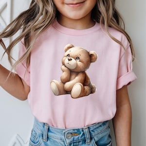 May include: A light pink t-shirt featuring a watercolor illustration of a teddy bear. The teddy bear is brown with a cream-colored belly and paws, and is sitting with its chin resting on its hand. The shirt is worn by a person.