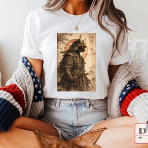 May include: White t-shirt featuring a detailed illustration of a samurai cat in full armor, set against a backdrop of a rising sun and Japanese text. The artwork is in shades of brown and black.