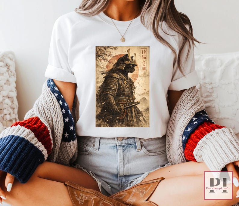 May include: White t-shirt featuring a detailed illustration of a samurai cat in full armor, set against a backdrop of a rising sun and Japanese text. The artwork is in shades of brown and black.
