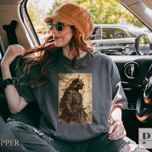 May include: A gray t-shirt featuring a vintage-style illustration of a samurai warrior with a red sun in the background. The design includes Japanese text and is printed on a comfortable, casual tee.