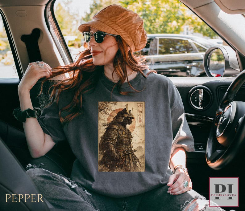 May include: A gray t-shirt featuring a vintage-style illustration of a samurai warrior with a red sun in the background. The design includes Japanese text and is printed on a comfortable, casual tee.