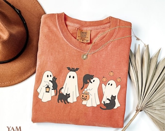 May include: Orange-red Comfort Colors t-shirt featuring a Halloween-themed graphic. The design includes cartoon ghosts, black cats, and pumpkins. The shirt has a round neckline and short sleeves. A gold necklace and a brown hat are also visible.