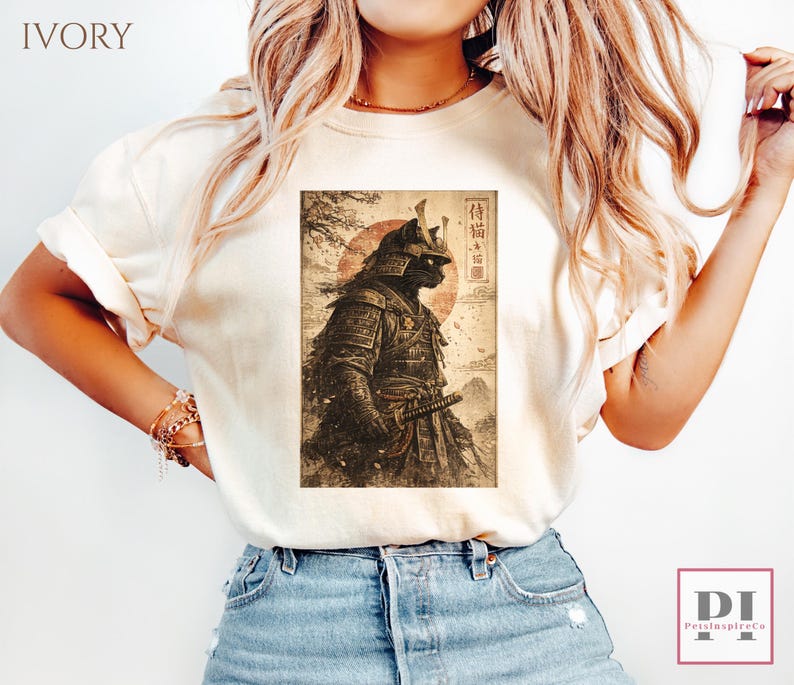 May include: Ivory-colored t-shirt featuring a graphic of a black cat in samurai armor, set against a red sun and landscape. The design includes Japanese text. The shirt is a casual wear item.