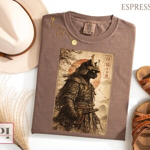 May include: Espresso-colored t-shirt featuring a graphic of a samurai cat with Japanese text. The shirt is surrounded by a white hat, gold necklace, and brown sandals. The shirt is a Comfort Colors brand, size XL-TG-EG.