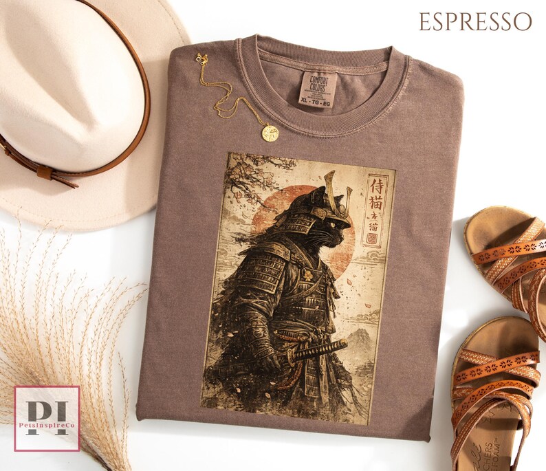 May include: Espresso-colored t-shirt featuring a graphic of a samurai cat with Japanese text. The shirt is surrounded by a white hat, gold necklace, and brown sandals. The shirt is a Comfort Colors brand, size XL-TG-EG.