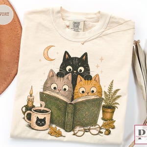 Puede incluir: Una camiseta color marfil con un estampado gráfico de tres gatos leyendo un libro verde abierto. El diseño incluye una luna creciente, una vela encendida, una taza con cara de gato, un helecho en maceta y gafas redondas. Etiqueta Comfort Colors.
