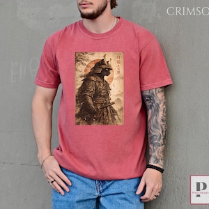 May include: Crimson-colored t-shirt featuring a detailed illustration of a samurai cat in traditional armor, holding a katana. The artwork is set against a backdrop of a rising sun and Japanese text. The shirt is made of a soft material.