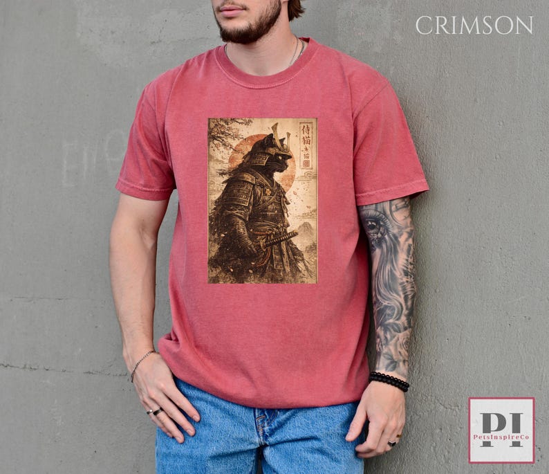 May include: Crimson-colored t-shirt featuring a detailed illustration of a samurai cat in traditional armor, holding a katana. The artwork is set against a backdrop of a rising sun and Japanese text. The shirt is made of a soft material.
