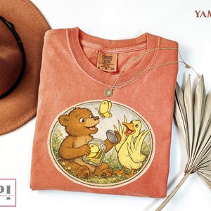 May include: A coral-colored t-shirt featuring a whimsical illustration of a bear, a duck, and a butterfly. The bear holds a jar with a duckling inside. The shirt has a crew neck and a Comfort Colors tag.
