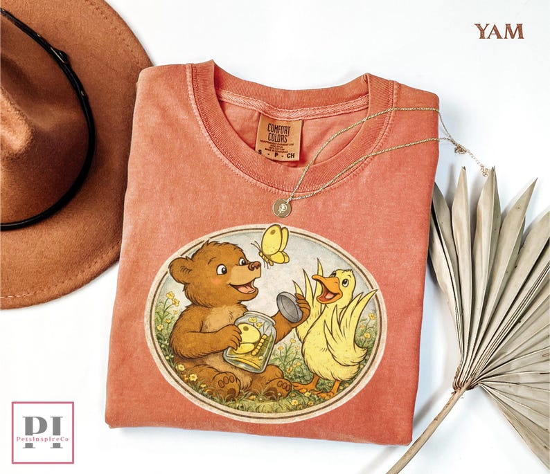 May include: A coral-colored t-shirt featuring a whimsical illustration of a bear, a duck, and a butterfly. The bear holds a jar with a duckling inside. The shirt has a crew neck and a Comfort Colors tag.