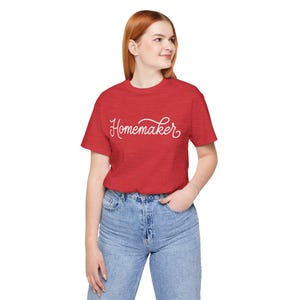 May include: A red short-sleeved t-shirt featuring the word Homemaker written in white cursive script across the chest. The shirt has a crew neckline and a heathered texture, paired with light blue denim jeans with a button and zipper fly.