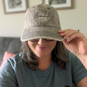 May include: A gray baseball cap with the embroidered text "Titus 2 Woman" in white script. The cap has a curved brim and a distressed, vintage look. The cap is being held by a person.