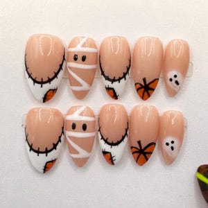 May include: A set of ten almond-shaped press-on nails featuring a nude base with Halloween-themed designs including white mummy bandages, black stitched lines, orange pumpkins, and small white ghosts with black eyes on a white background.