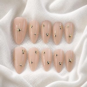 Op de afbeelding: Een set van tien amandelvormige press-on nagels in een nude beige kleur. Elke nagel is voorzien van gouden metallic steraccenten en kleine gouden metallic drie-cirkel karakterpictogrammen in diverse patronen op het gladde oppervlak.