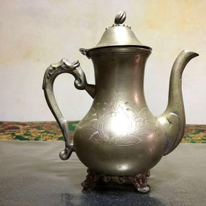 Peut inclure: Théière ancienne de couleur argentée avec une anse et un bec décoratifs. La théière présente un motif floral orné sur le corps et repose sur une petite base à trois pieds. Le couvercle est surmonté d'un petit fleuron décoratif.