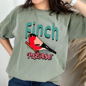 May include: Sage green t-shirt featuring a colorful illustration of a finch perched on a branch. The word "Finch" is in teal, and "PLEASE" is in pink. The design is centered on the front of the shirt.