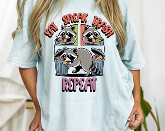 May include: Light blue t-shirt with a graphic of raccoons and trash cans. The text reads "EAT SNEAK TRASH REPEAT" in a colorful, playful font. The design features three raccoons, two in trash cans and one outside.