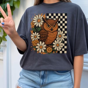 May include: Dark grey t-shirt with a brown owl illustration, daisies, and a checkered pattern. The owl has orange and brown feathers, and the daisies have white petals and orange centres. The shirt is a casual style.