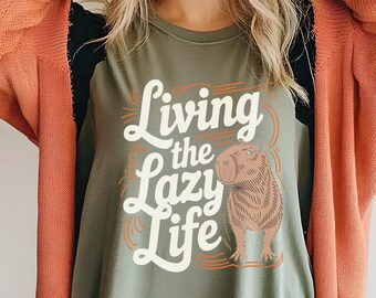 May include: Olive green t-shirt with the phrase "Living the Lazy Life" in white script and a brown capybara illustration. The shirt has a relaxed fit and is made of soft material.