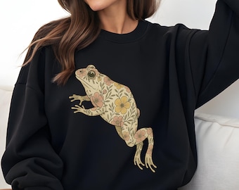 May include: Black sweatshirt featuring a floral frog design in beige, yellow, and pink. The frog illustration is detailed with a vintage aesthetic. The sweatshirt is a casual, comfortable style.
