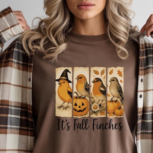 May include: A brown t-shirt with a design of four finches, each on a separate panel, with pumpkins and autumn leaves. One finch wears a witch's hat. The text "It's Fall Finches" is printed below.