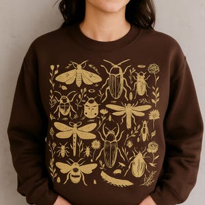 May include: A brown sweatshirt with a gold insect and floral pattern. The design features various insects, including beetles, dragonflies, and moths, alongside floral elements. The design is centrally placed on the front of the sweatshirt.