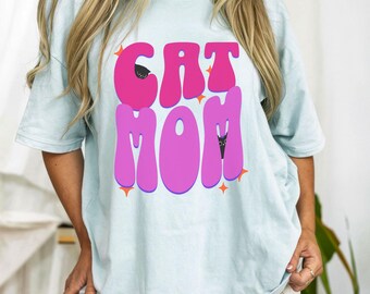 May include: Light blue t-shirt with the text "CAT MOM" in pink bubble letters. The design includes small black cat silhouettes within the letters and orange star accents. The shirt is a casual, oversized style.