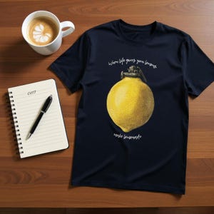 Peut inclure: T-shirt bleu marine avec un citron jaune en forme de grenade et le texte "When life gives you lemons, make lemonade." Une tasse de café et un bloc-notes avec un stylo sont sur une surface en bois.