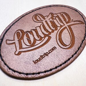 Op de afbeelding: Een bruin leren patch met het woord "Loudtrip" in een gestileerd lettertype en de website "loudtrip.com" eronder.